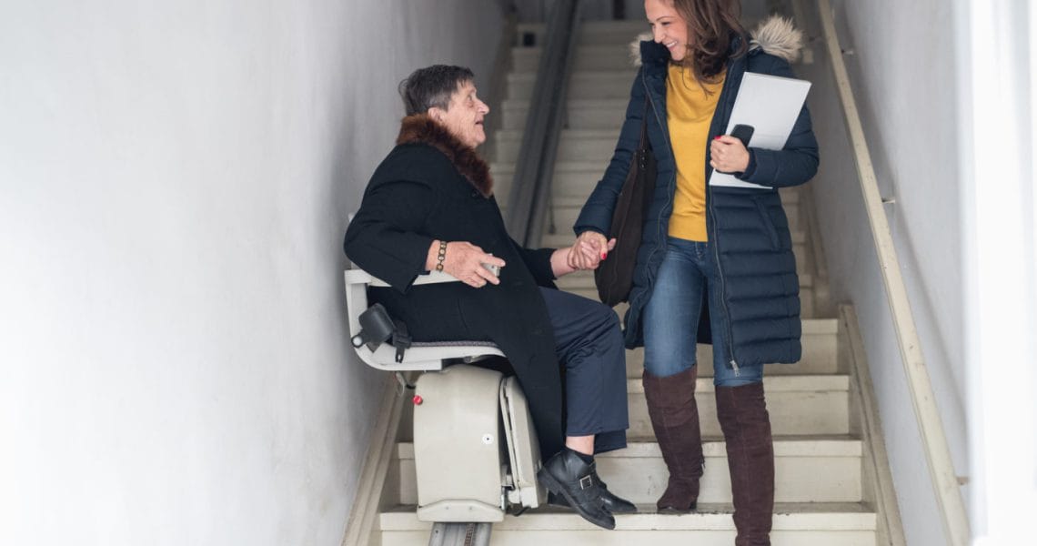 A young woman smiling at an elderly woman seated on a stair lift indoors.