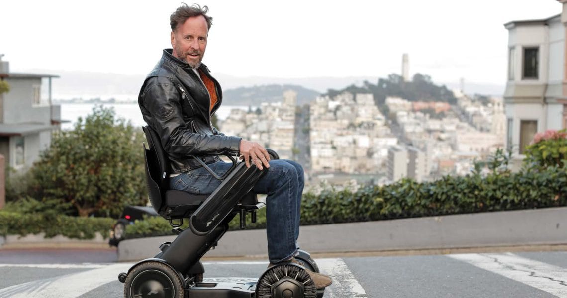Man sitting on a compact electric vehicle on an inclined street with cityscape in the background.