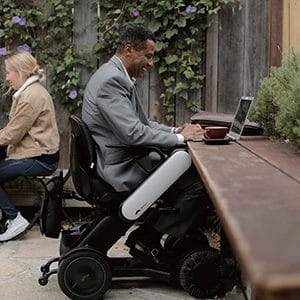 Man in a motorized wheelchair working on a laptop at an outdoor table.